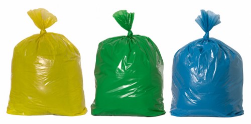 Three large plastic rubbish sacks are positioned side by side outdoors, with the yellow sack on the left, the green in the center, and the blue on the right. Each sack is filled, with visible creases and a glossy finish reflecting light, and tied at the top with the plastic gathered and knotted. The environment appears to be a paved driveway or external ground surface, with the sacks standing upright on the pavement. The background is minimal and out of focus, emphasizing the sacks' bright colours and wrinkled textures. The scene relates to waste collection or disposal services, typical of commercial waste removal operations that a company like Commercial Waste Highbury might provide in the Highbury area, with this image illustrating common rubbish bag contents and packaging as part of the recycling or disposal process.