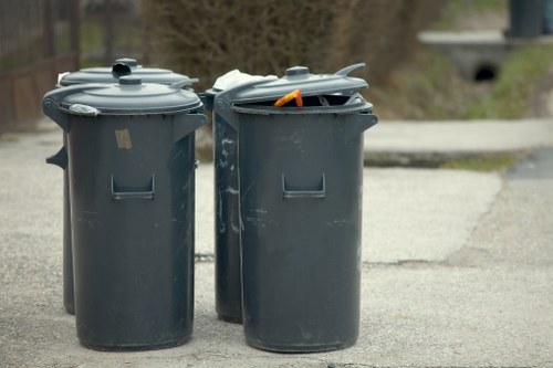Two large cylindrical black waste bins with tightly fitting lids are positioned on a paved outdoor surface, likely a driveway or street in Highbury. The bin on the right has an orange object, possibly part of a clothing item or other material, protruding from its open lid, while the bin on the left has a sticker or label on its lid. Both bins are made of sturdy plastic with a matte finish and rectangular indentations on the front for structural reinforcement. They are situated side by side, with the background showing blurred natural elements such as trees and a low wall, indicating an urban or suburban environment. The scene is illuminated by natural daylight, emphasizing the solid materials and functional design of the waste containers. This setup aligns with typical rubbish disposal arrangements in residential areas served by Commercial Waste Highbury, emphasizing their role in household or commercial waste collection in the Highbury postcode area.