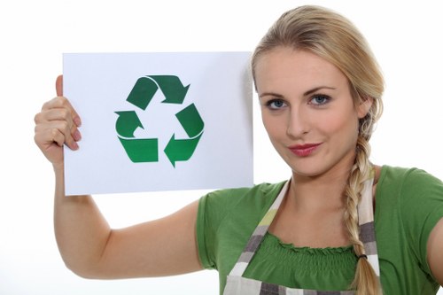 A young woman with blonde hair styled in a side braid, wearing a green top and a striped apron, holds a white sign with a large green recycling symbol composed of three arrows forming a triangle. She is standing against a plain white background. The image suggests themes of waste management and recycling awareness, reflecting environmentally responsible practices often associated with commercial rubbish removal services. In this context, the woman’s expression is neutral and professional, deliberately emphasizing the recycling symbol to support waste reduction initiatives. The focus on the recycling icon emphasizes environmental sustainability, which aligns with services offered by companies such as Commercial Waste Highbury in London, within the N19 postcode area. Her casual attire and clear depiction of the recycling symbol highlight a community-oriented approach to rubbish disposal and waste management efforts typically seen in local commercial rubbish removal activities related to Highbury and nearby areas.