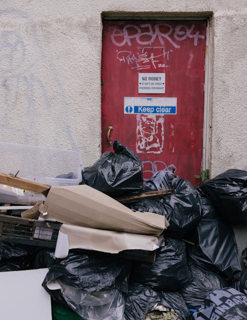 The image shows four blue rubbish bags placed on a concrete sidewalk in front of a grey metal door with a black lever handle, set within an entrance to a building. The building’s exterior wall is te