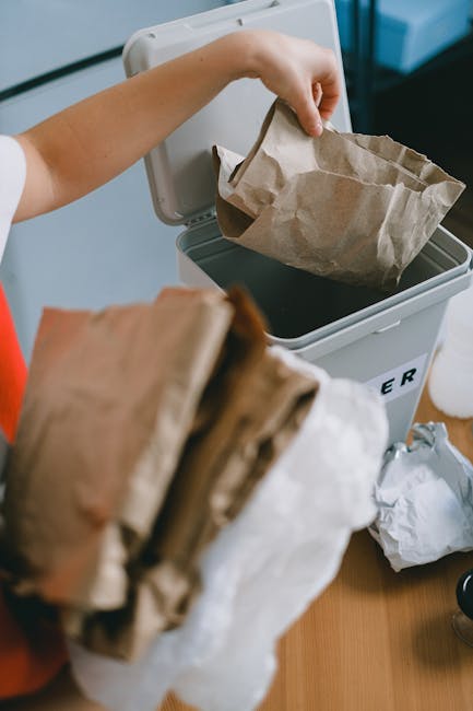 A person's hand is seen placing a crumpled brown paper bag into a white plastic rubbish bin with the label 'RUBBISH' on it. The bin is positioned on a wooden surface within an indoor setting, possibly