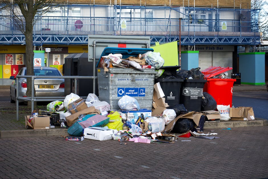 A person's hand is seen placing a crumpled brown paper bag into a white plastic rubbish bin with the label 'RUBBISH' on it. The bin is positioned on a wooden surface within an indoor setting, possibly