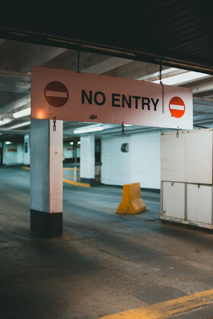 An indoor parking garage scene featuring a large rectangular sign suspended from the ceiling, displaying a red circle with a white horizontal line and the words 'NO ENTRY' in bold black letters, indic